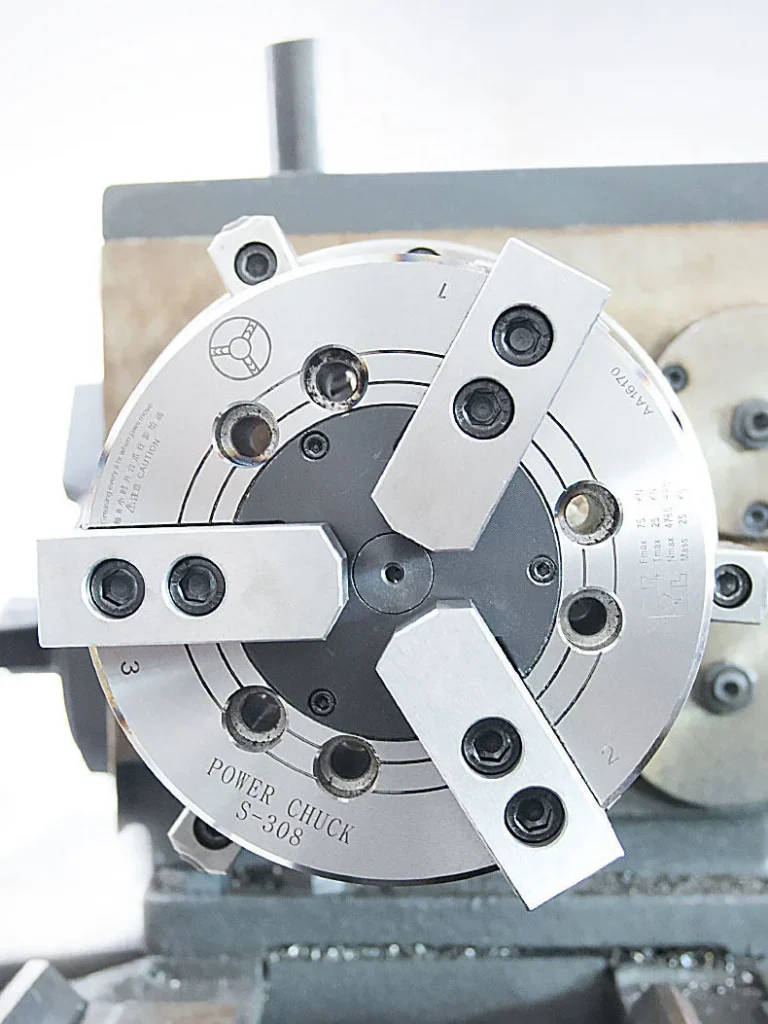Close-up photograph of a 3-jaw lathe chuck with all three jaws installed, showing jaw positioning, gripping surface, and mounting details on the chuck body.
